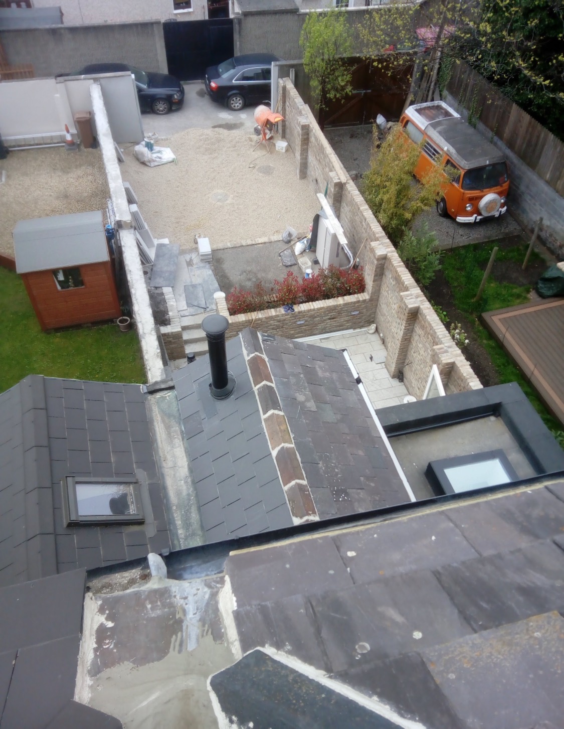 Widok z góry na remontowany dach z oknem, z fragmentem podwórka z budynkiem gospodarczym, samochodem i osobą pracującą przy betoniarce w tle.