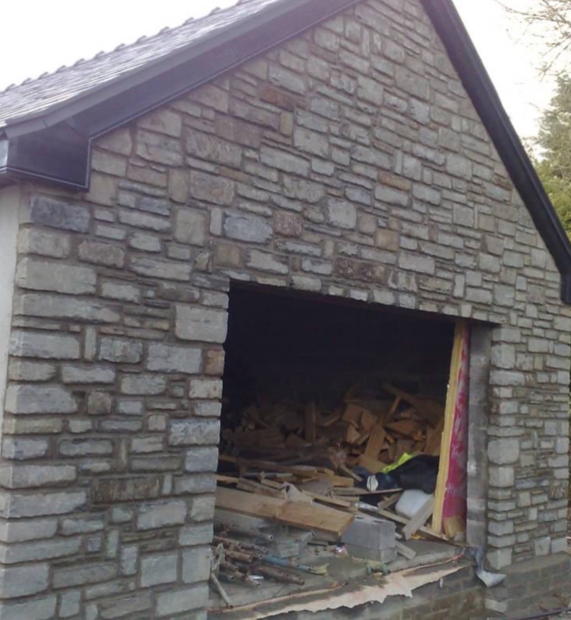 Budynek garażowy w trakcie budowy, ściany wyłożone kamieniem, widoczne materiały budowlane wewnątrz, surowy stan wykończenia.