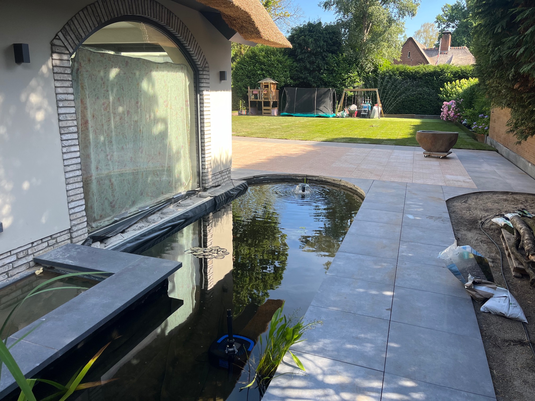 Oczko wodne z fontanną i kamienną obudową, otoczone szarymi płytami chodnikowymi. W tle trawnik, plac zabaw i dom z trzcinowym dachem.