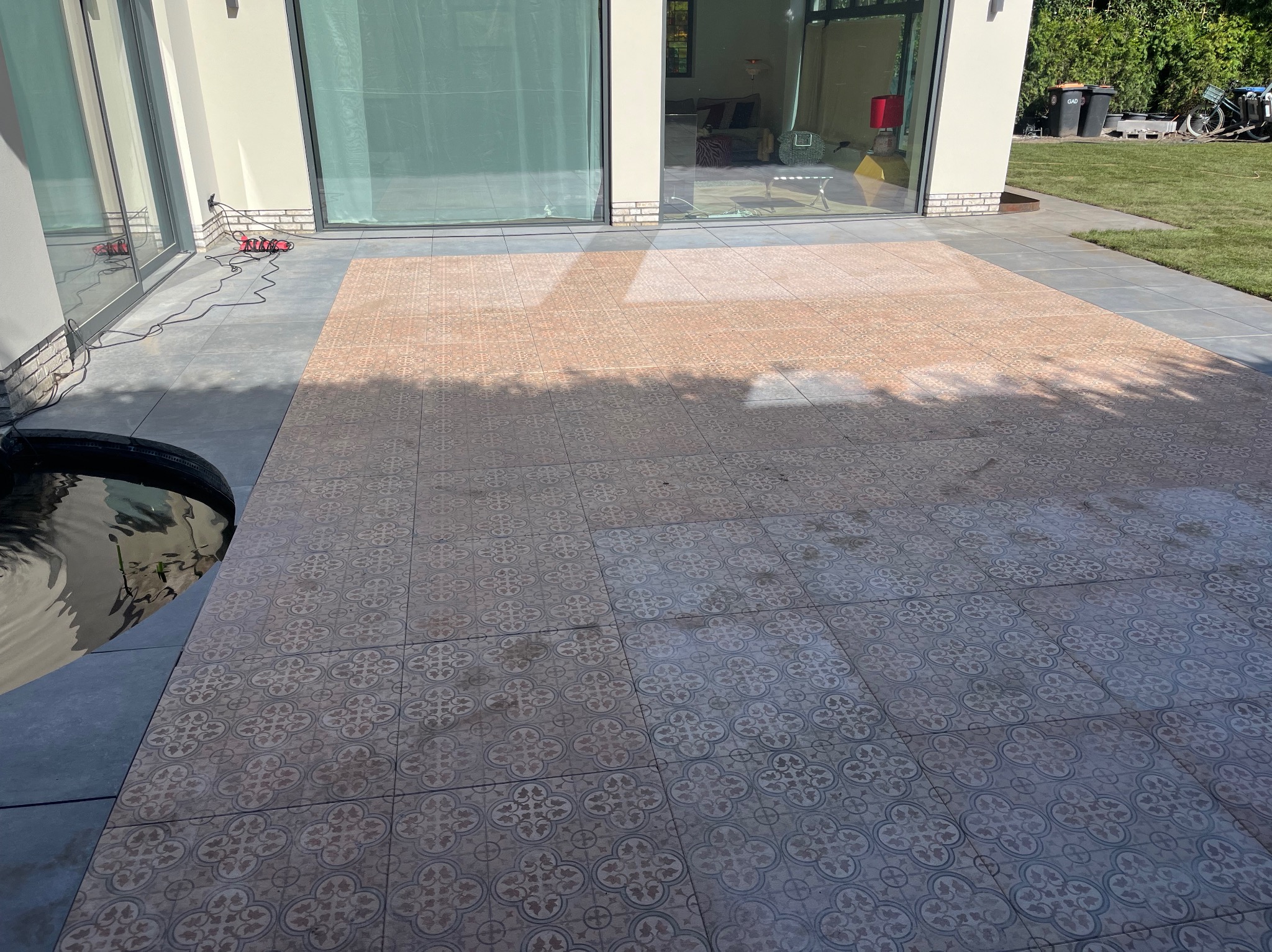 Wykończony taras z wzorzystą terakotą, przylegający do domu z dużymi przesuwnymi oknami. Widoczny fragment oczka wodnego i trawnik.