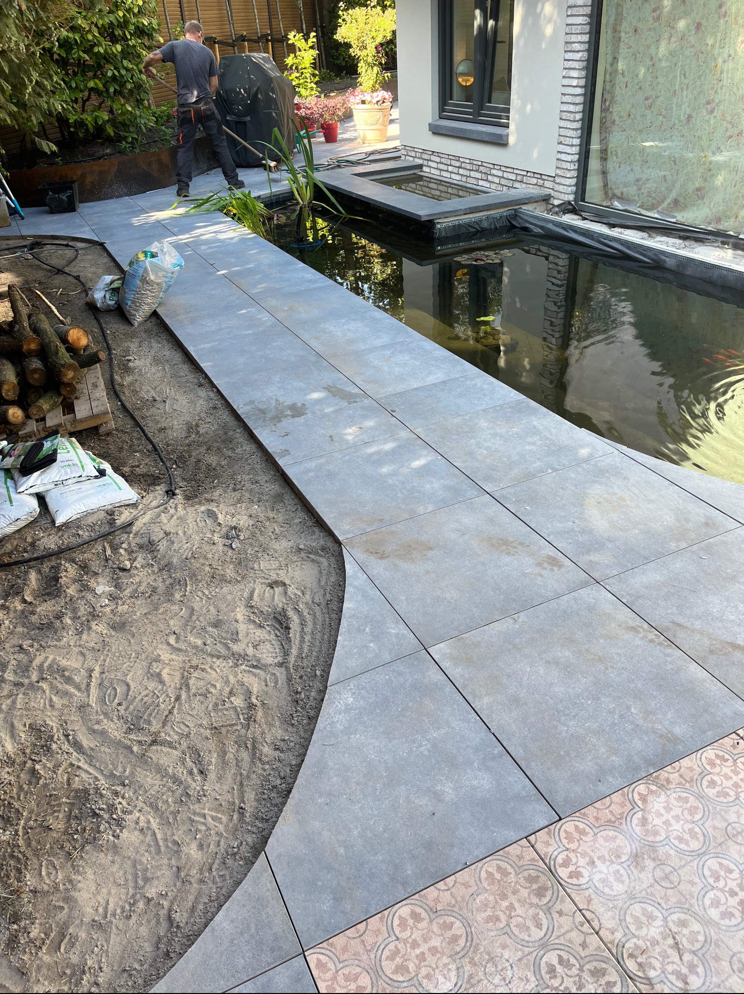 Układanie szarej, kwadratowej kostki brukowej wokół oczka wodnego w ogrodzie. Widoczny pracownik w tle i worki z materiałami budowlanymi.