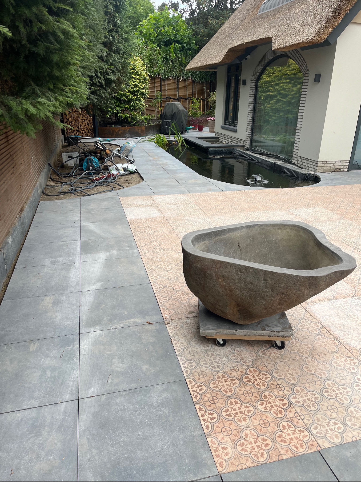 Układanie płytek na tarasie z dekoracyjnym zbiornikiem wodnym i kamienną donicą. Widoczne narzędzia i materiały budowlane. Prace wykończeniowe w ogrodzie.