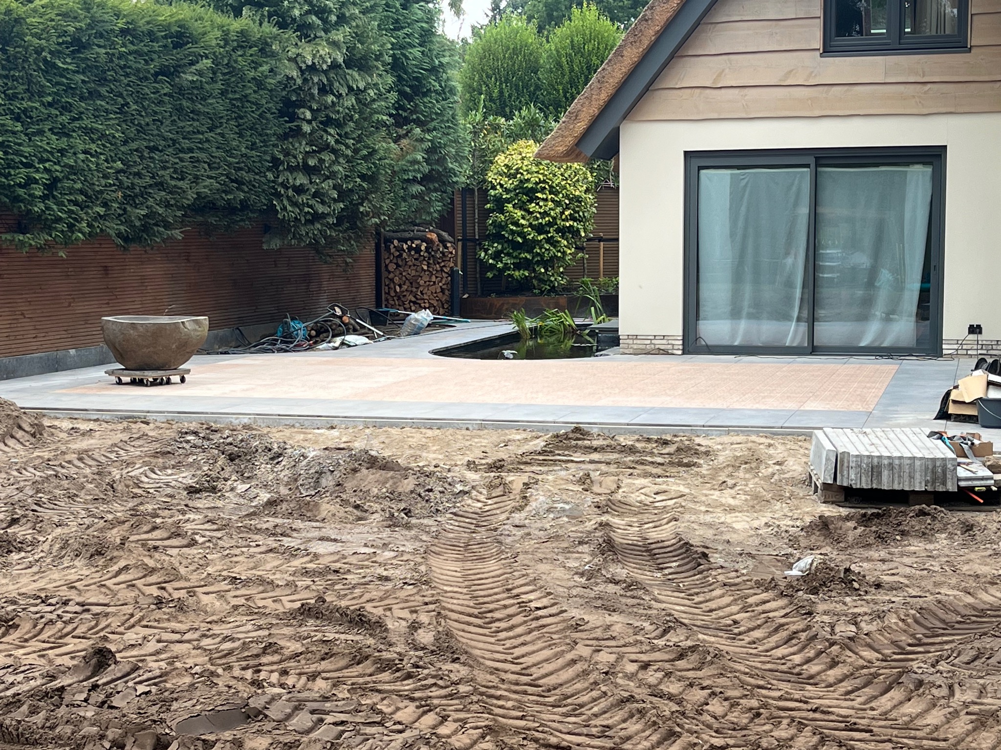 Układanie kostki brukowej na posesji z oczkiem wodnym i zielenią. Widoczne ślady opon na ziemi, przygotowane płyty i fragment domu z przesuwnymi drzwiami.