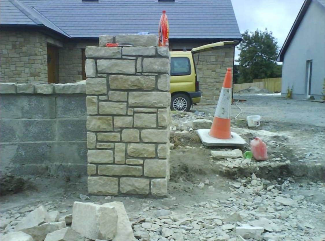 Wzniesiony kamienny słupek ogrodzeniowy z nieregularnych bloków, z żółtym vanem i pachołkiem drogowym w tle, na placu budowy.