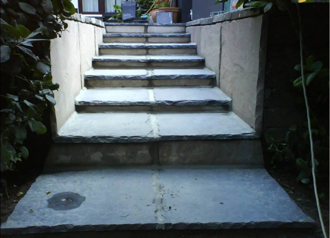 Kamienne schody zewnętrzne z roślinnością po bokach, prowadzące do domu. Widoczne szare płyty kamienne i jasne spoiny. Ujęcie z dołu, podkreślające solidność konstrukcji.