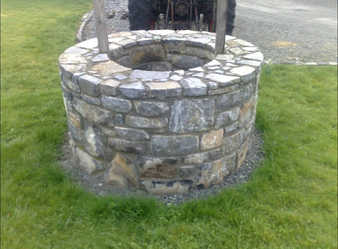 Studnia z kamienia polnego na trawniku, z drewnianymi słupkami i traktorem w tle. Ręczna robota, nieregularne kamienie, naturalny wygląd.