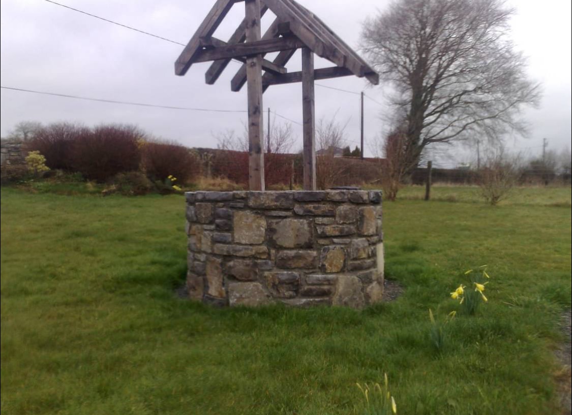 Kamienna studnia z drewnianą konstrukcją na zielonym trawniku, w tle drzewa i krzewy. Element architektury ogrodowej w naturalnym otoczeniu.