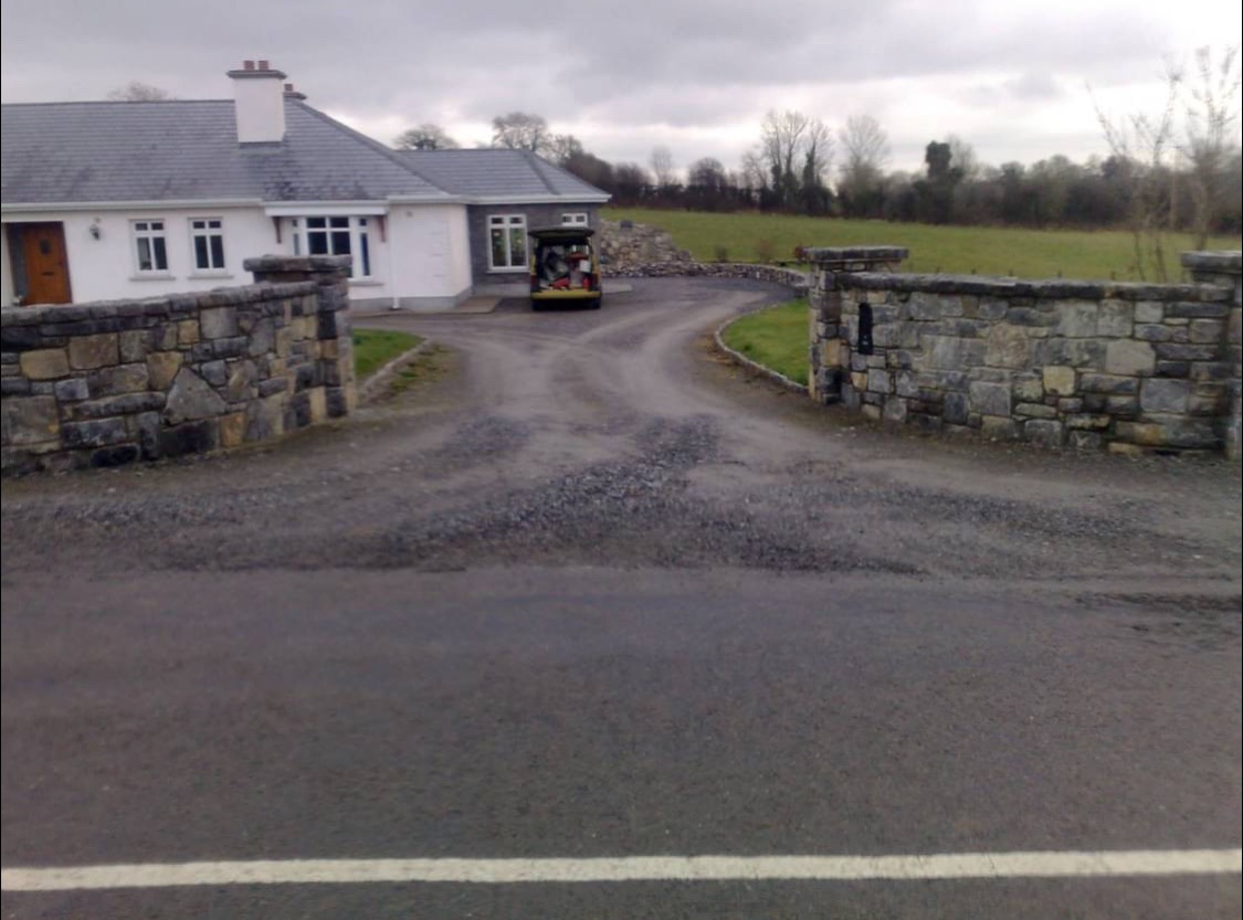 Dom jednorodzinny z szarym dachem i kamiennym ogrodzeniem, widok z drogi. Na podjeździe stoi żółty samochód dostawczy.