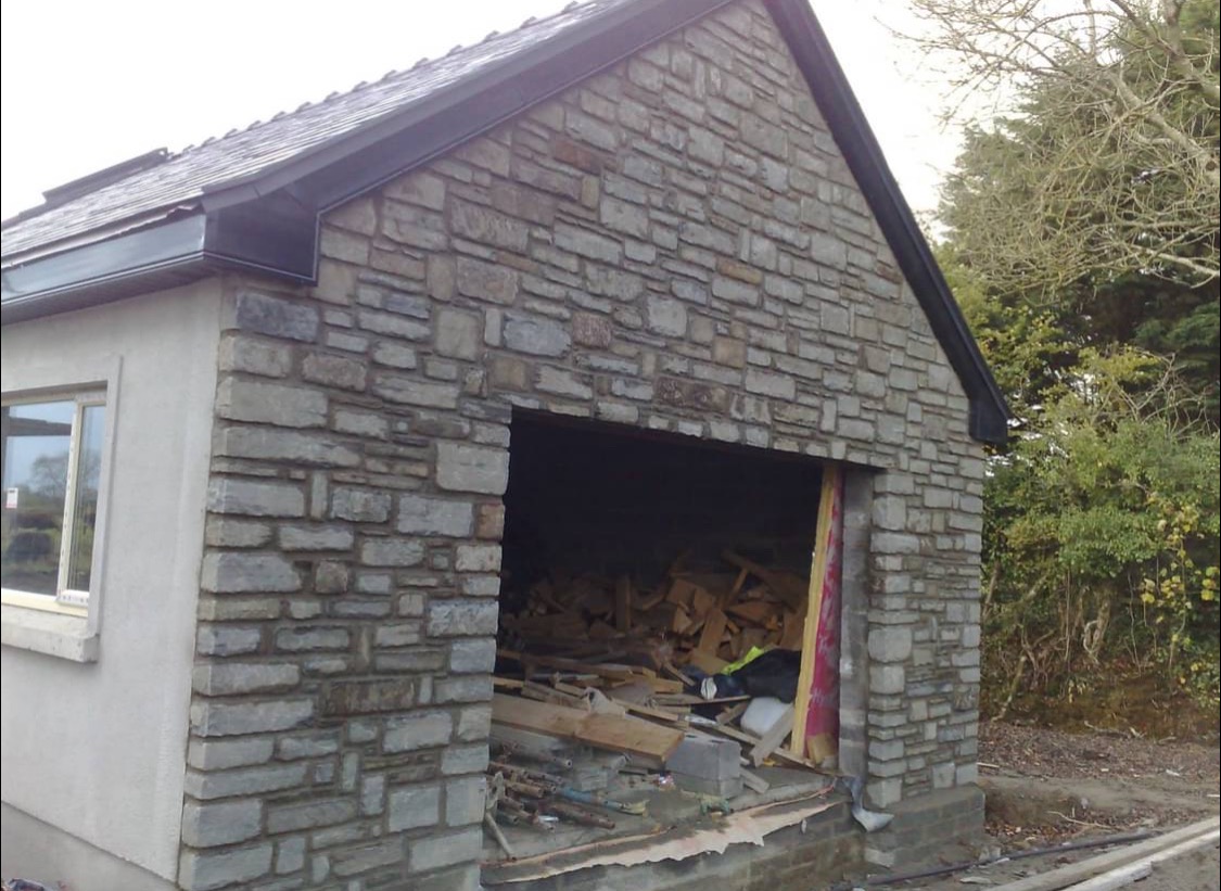 Garaż z elewacją z kamienia, widoczne materiały budowlane wewnątrz. Część ściany otynkowana na biało, okno. Dach pokryty dachówką.