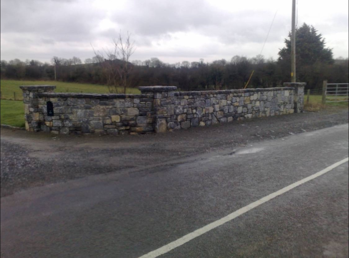 Kamienny mur oporowy z filarami, oddzielający drogę od zielonego terenu. Widoczne nieregularne kamienie i szara kolorystyka. Pochmurne niebo w tle.