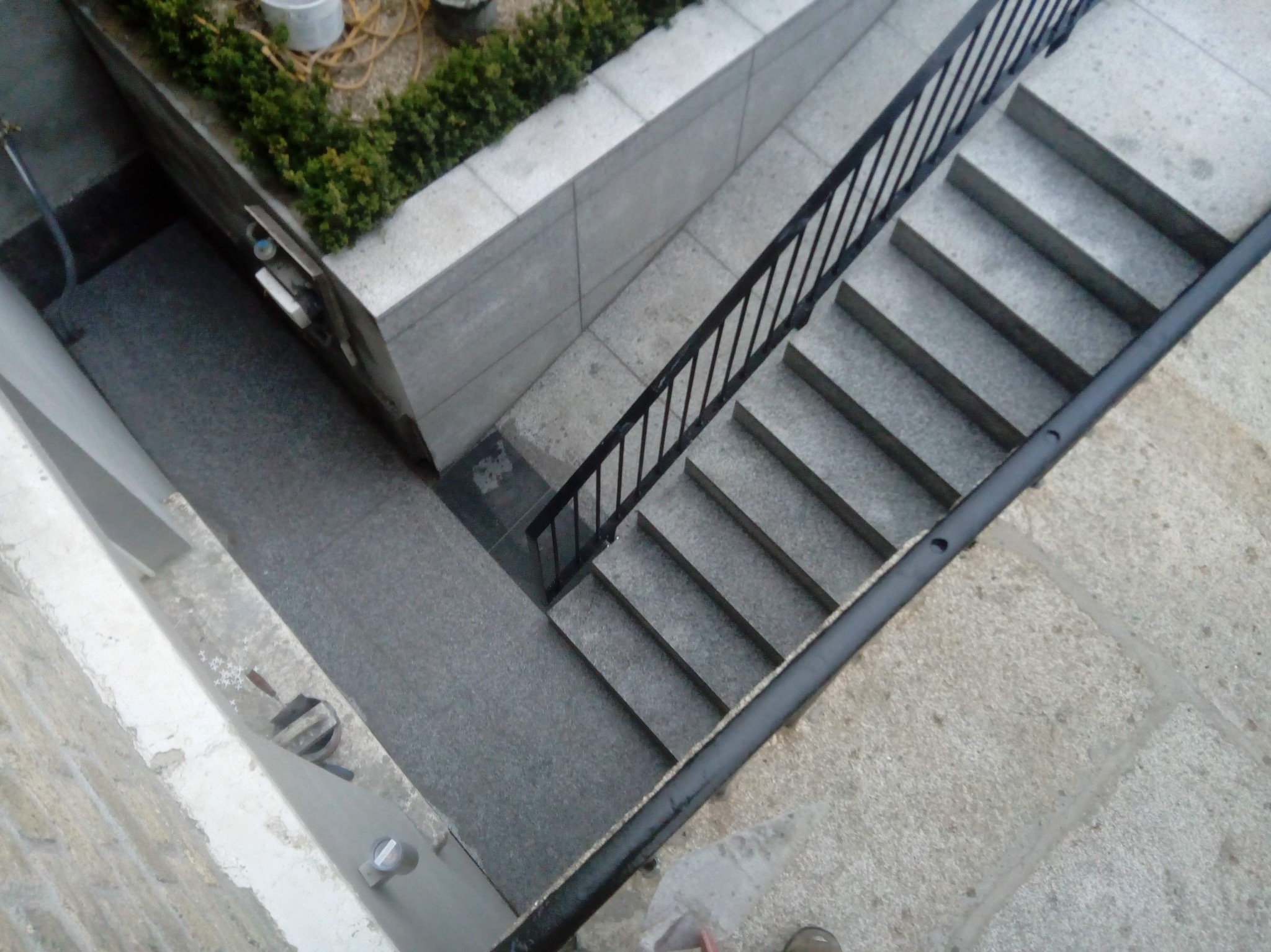 Schody zewnętrzne z szarej kostki brukowej, zabezpieczone czarną metalową barierką. Widok z góry, z fragmentem zieleni w tle. Minimalistyczny design.