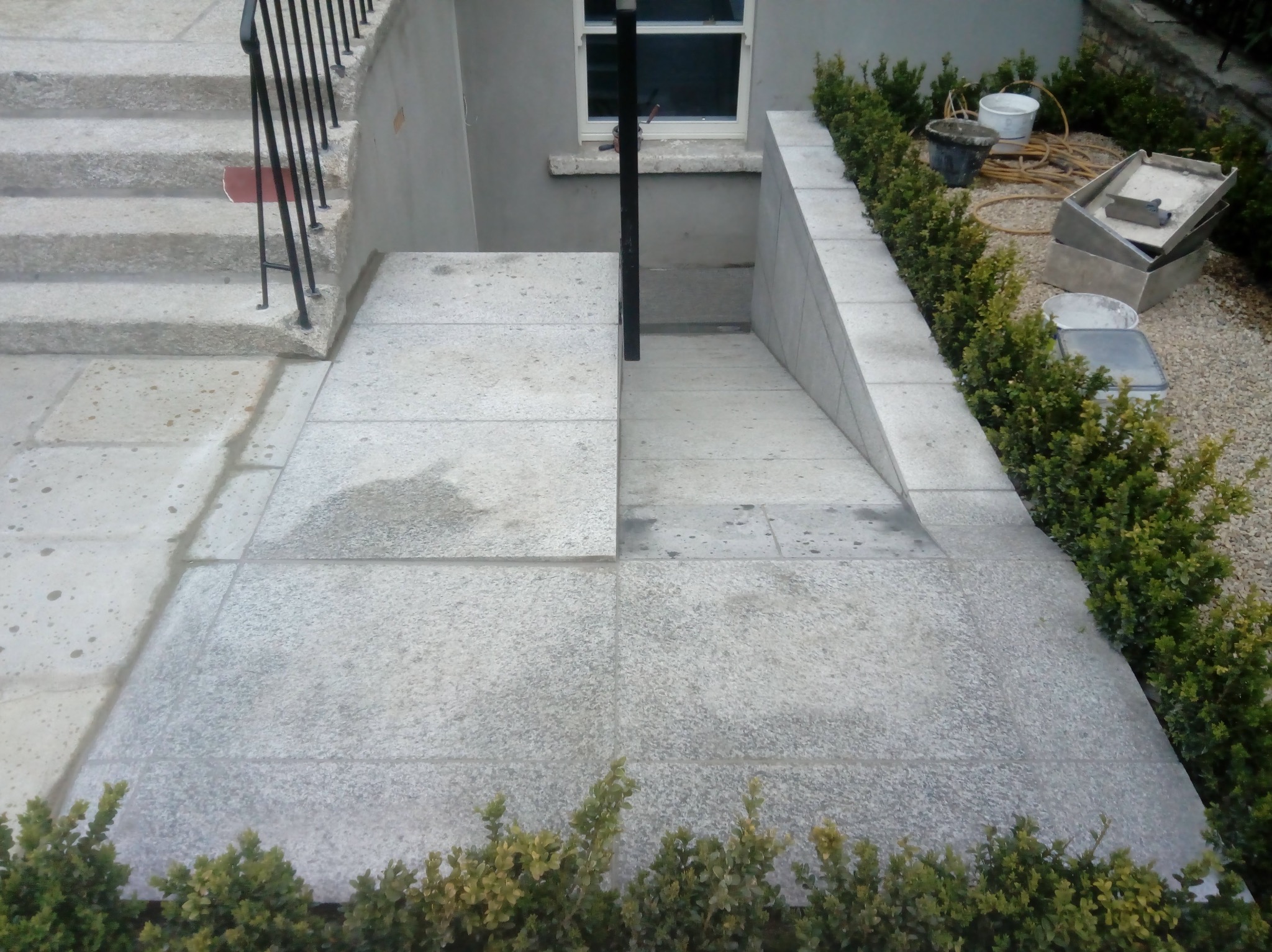 Wykończone kamienne schody zewnętrzne z balustradą i płytkami, prowadzące do piwnicy. Widoczne narzędzia i roślinność. Szare odcienie kamienia i zieleni.