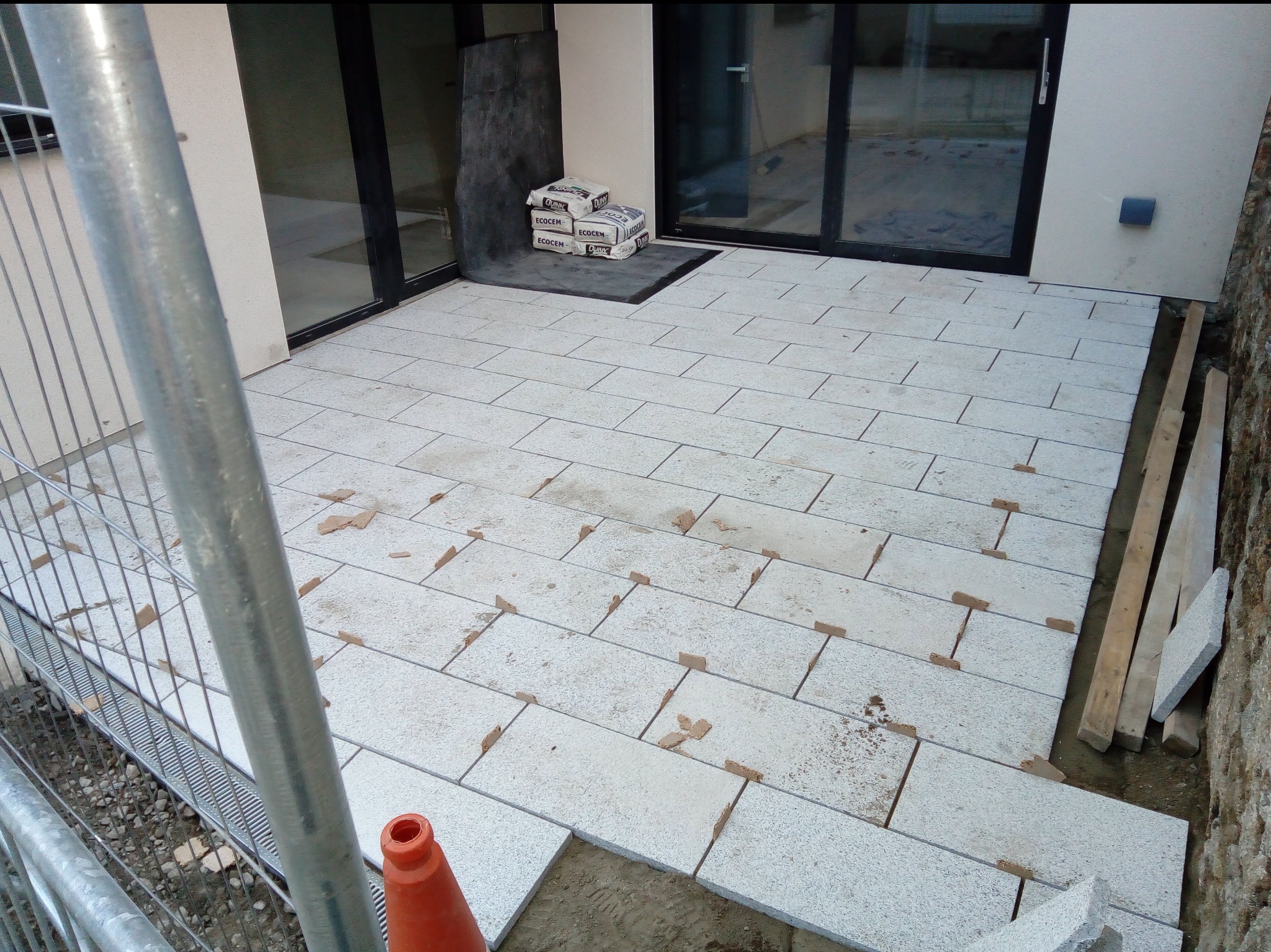 Układanie jasnych płyt na tarasie zewnętrznym, widoczne materiały budowlane i zabezpieczenia. Prace w toku, częściowo ułożona nawierzchnia.