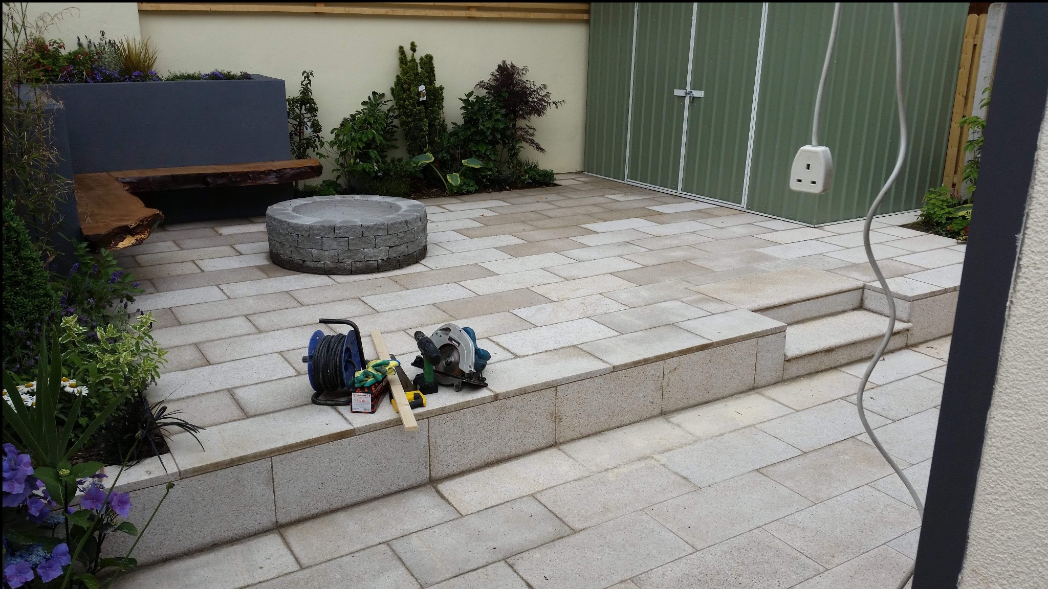 Wykończony taras z kamiennej kostki w Aleksandrowie, z narzędziami pracy na pierwszym planie. Widoczny fragment ogrodu i schowek narzędziowy w tle.