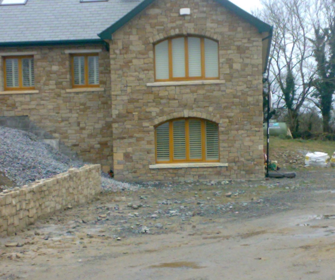 Dom z elewacją z kamienia naturalnego, z widocznym fragmentem muru oporowego i stertą gruzu, sugerujący prace wykończeniowe.