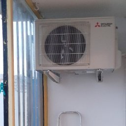 FOGTECH - Biała jednostka zewnętrzna klimatyzatora Mitsubishi Electric zamontowana na balkonie, widoczne uchwyty montażowe i fragment drabiny, z widokiem na budynki przez pionowe żaluzje.