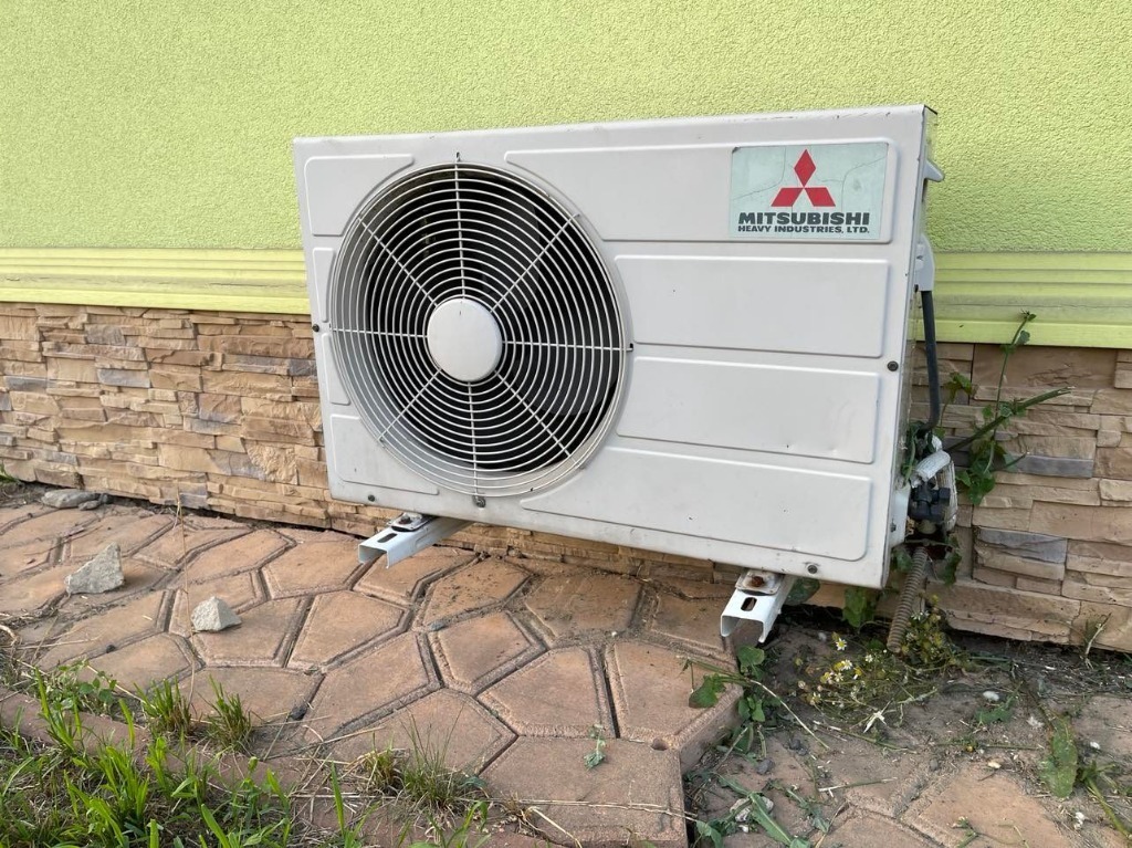 Zewnętrzna jednostka klimatyzatora Mitsubishi Heavy Industries zamontowana na elewacji domu z zielonym tynkiem i kamienną podmurówką, widoczne ślady użytkowania i roślinność wokół.