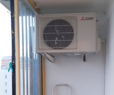 Biała jednostka zewnętrzna klimatyzatora Mitsubishi Electric zamontowana na balkonie, widoczne uchwyty montażowe i fragment drabiny, z widokiem na budynki przez pionowe żaluzje.