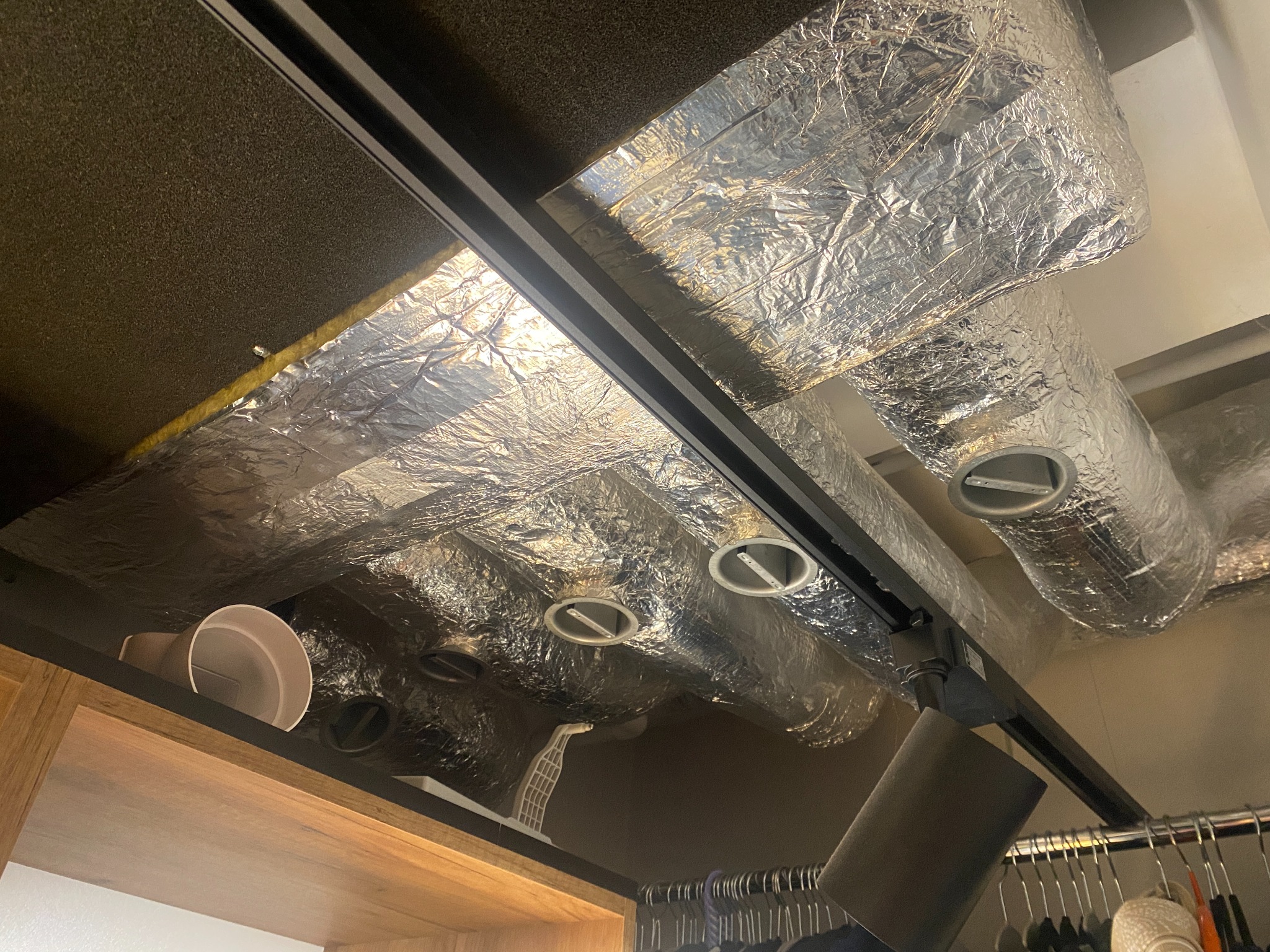 Widok od dołu na izolowane kanały wentylacyjne z okrągłymi wylotami, zamontowane pod sufitem w garderobie z widocznymi ubraniami na wieszakach.