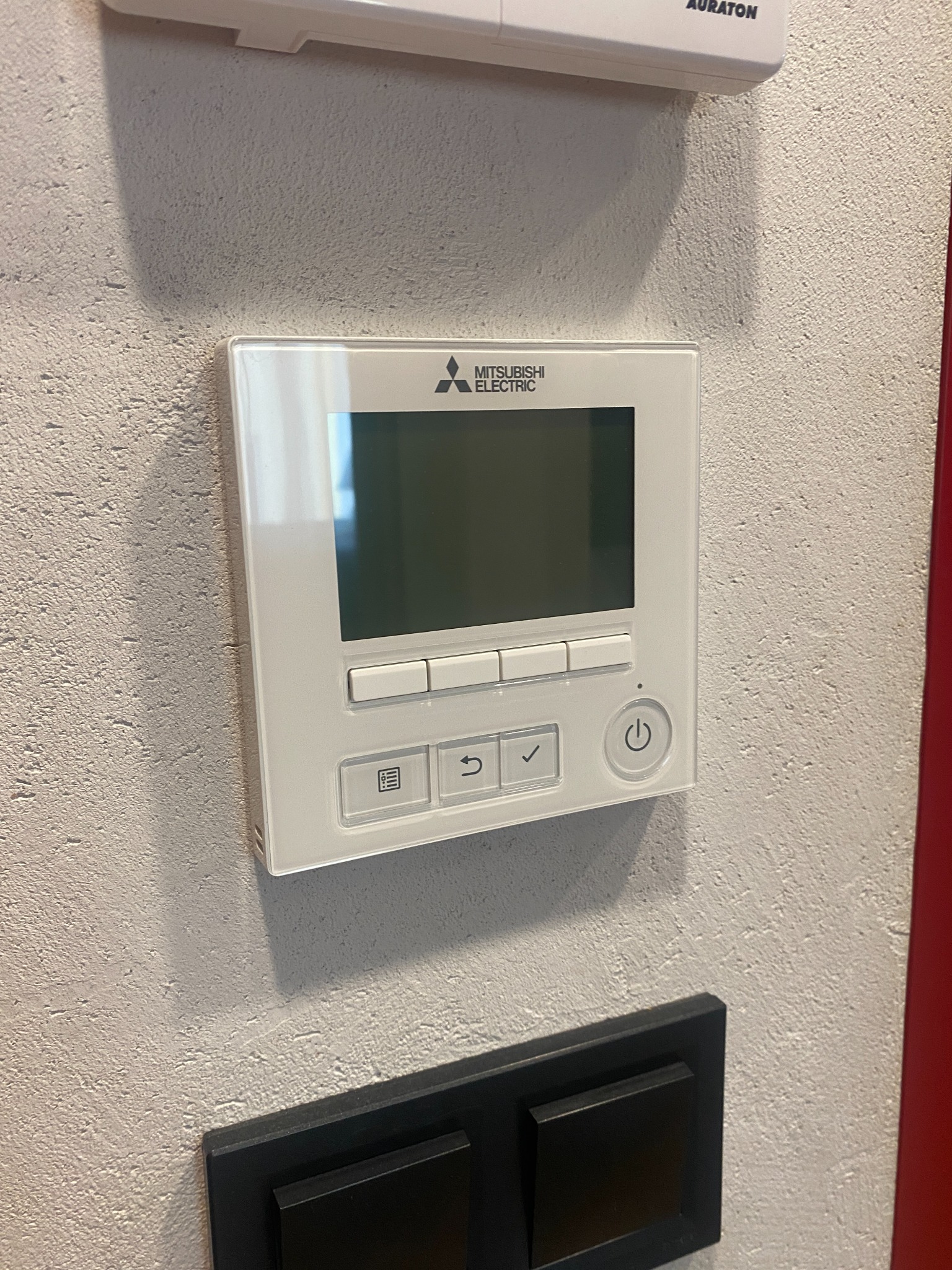 Biały panel sterowania klimatyzacją Mitsubishi Electric zamontowany na szarej, teksturowanej ścianie, poniżej czarny włącznik światła, widoczny fragment urządzenia Auraton na górze.