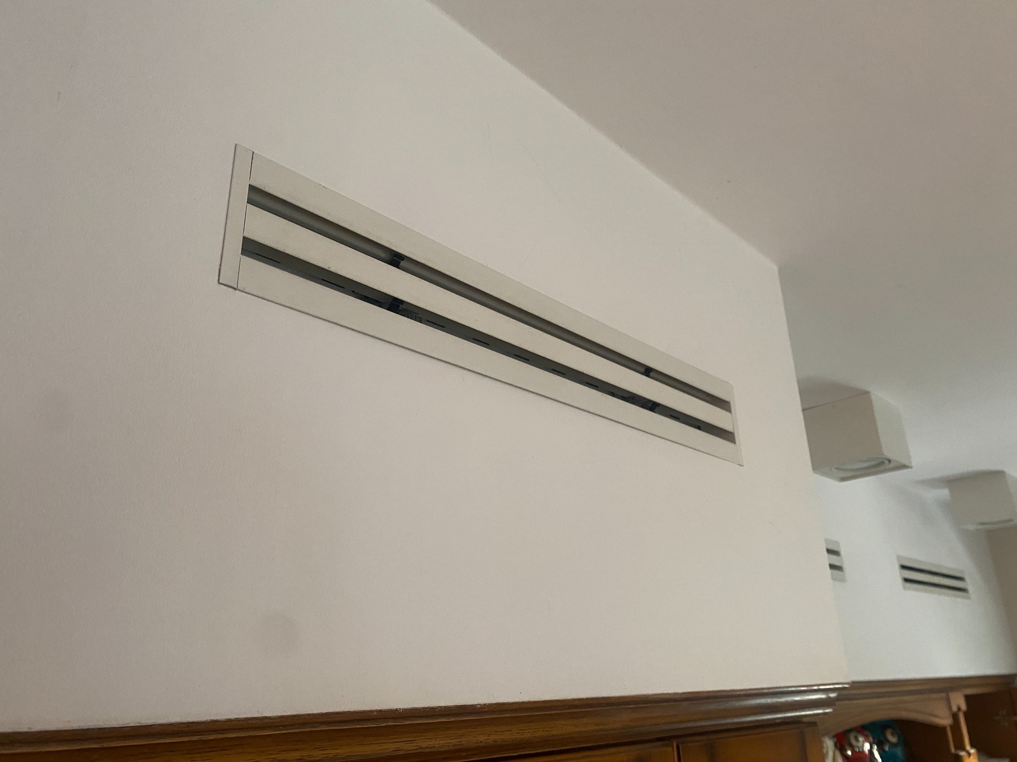 Biała, prostokątna kratka wentylacyjna z trzema poziomymi szczelinami, zamontowana na białej ścianie w pomieszczeniu z drewnianą boazerią i widocznymi elementami instalacji wentylacyjnej na suficie.