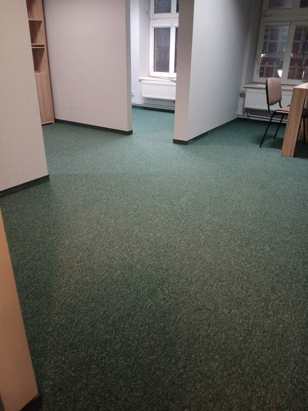 Wnętrze biurowe z zieloną wykładziną dywanową, widok na sąsiednie pomieszczenia przez otwory w ścianach, grzejniki pod oknami.