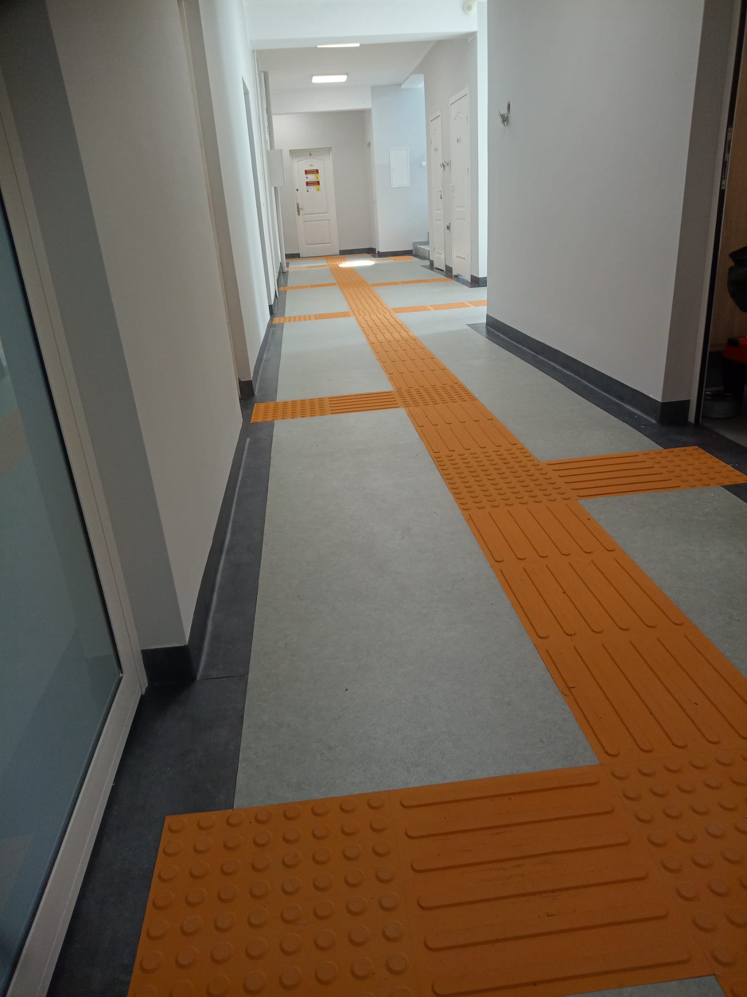 Korytarz z szarą posadzką i pomarańczowymi, dotykowymi pasami nawigacyjnymi dla osób niedowidzących, z widocznymi wypukłościami i liniami, prowadzącymi przez całą długość korytarza.
