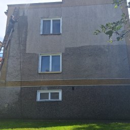 ARKFI Usługi Wysokościowe A. NOWAK, F. CYGANEK spółka cywilna - Pracownik na drabinie spryskuje elewację budynku, widoczna różnica w kolorze po umyciu fragmentu ściany, trzy okna na różnych poziomach, trawnik u dołu.