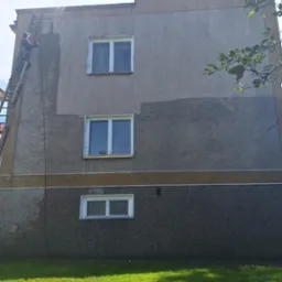 Pracownik na drabinie spryskuje elewację budynku, widoczna różnica w kolorze po umyciu fragmentu ściany, trzy okna na różnych poziomach, trawnik u dołu.