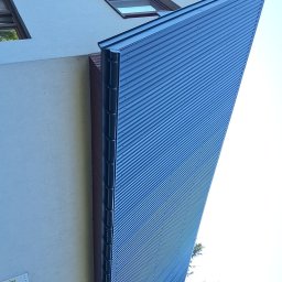 ARKFI Usługi Wysokościowe A. NOWAK, F. CYGANEK spółka cywilna - Fragment elewacji budynku z nową, niebieską blachą trapezową, widok z dołu, z widocznym fragmentem dachu i drzewa w tle.