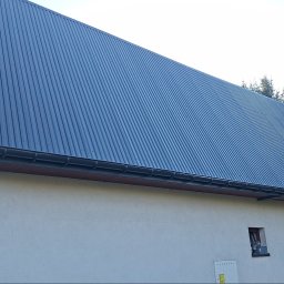 ARKFI Usługi Wysokościowe A. NOWAK, F. CYGANEK spółka cywilna - Dom jednorodzinny z nowym, ciemnoszarym dachem z blachy trapezowej i zamontowanymi rynnami. Widoczna elewacja w jasnym kolorze. W tle iglaste drzewa.