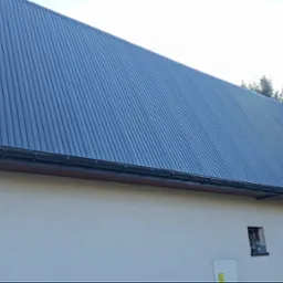 Dom jednorodzinny z nowym, ciemnoszarym dachem z blachy trapezowej i zamontowanymi rynnami. Widoczna elewacja w jasnym kolorze. W tle iglaste drzewa.