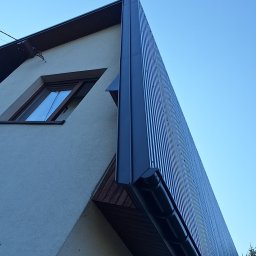 ARKFI Usługi Wysokościowe A. NOWAK, F. CYGANEK spółka cywilna - Fragment elewacji budynku z nowoczesnym, pionowo żłobkowanym, ciemnoszarym panelem elewacyjnym, widok z dołu na tle błękitnego nieba.