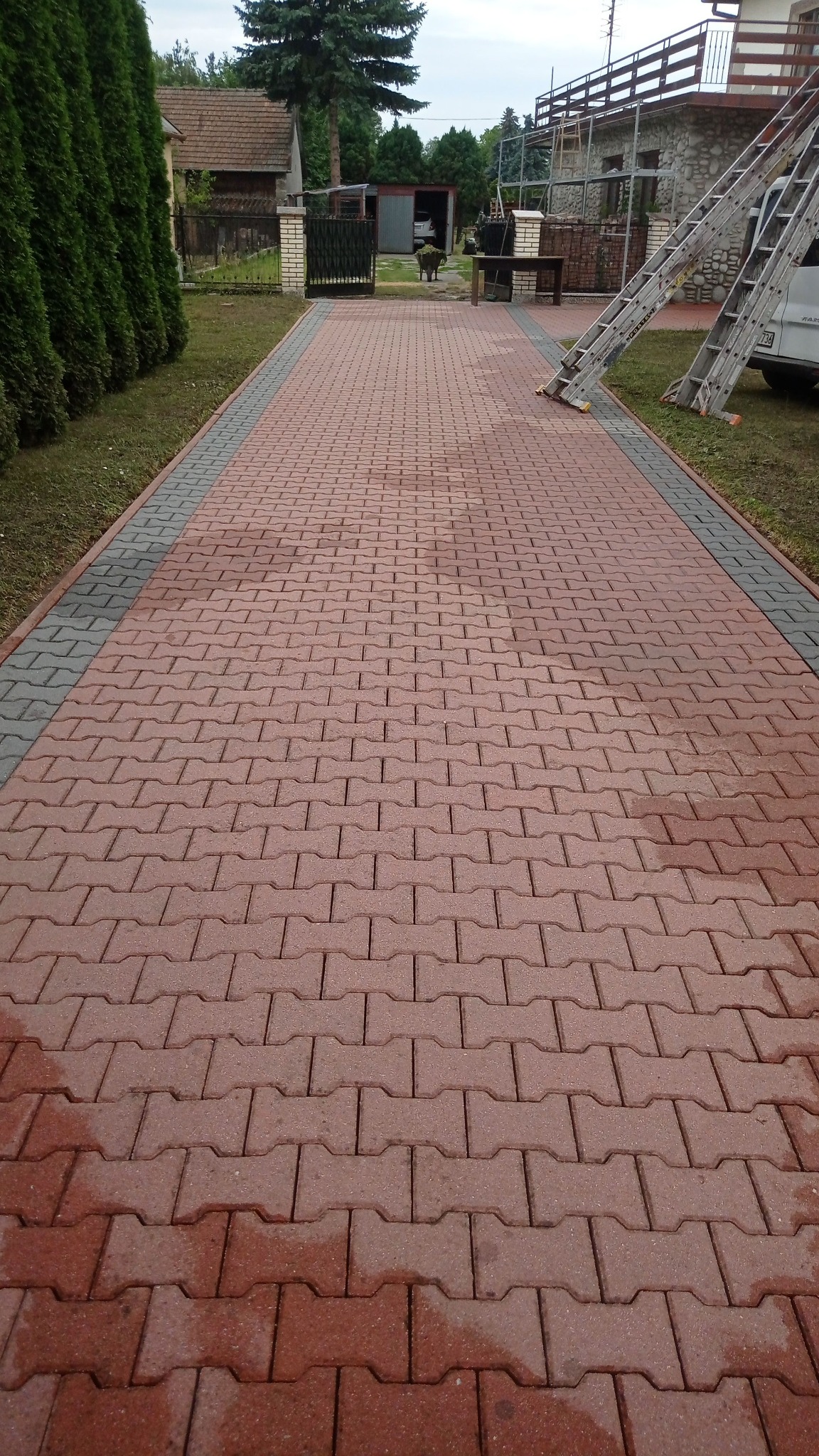 Czerwona kostka brukowa ułożona na podjeździe, widoczne ślady wilgoci, w tle dom w trakcie prac remontowych, oparty o elewację drabina.