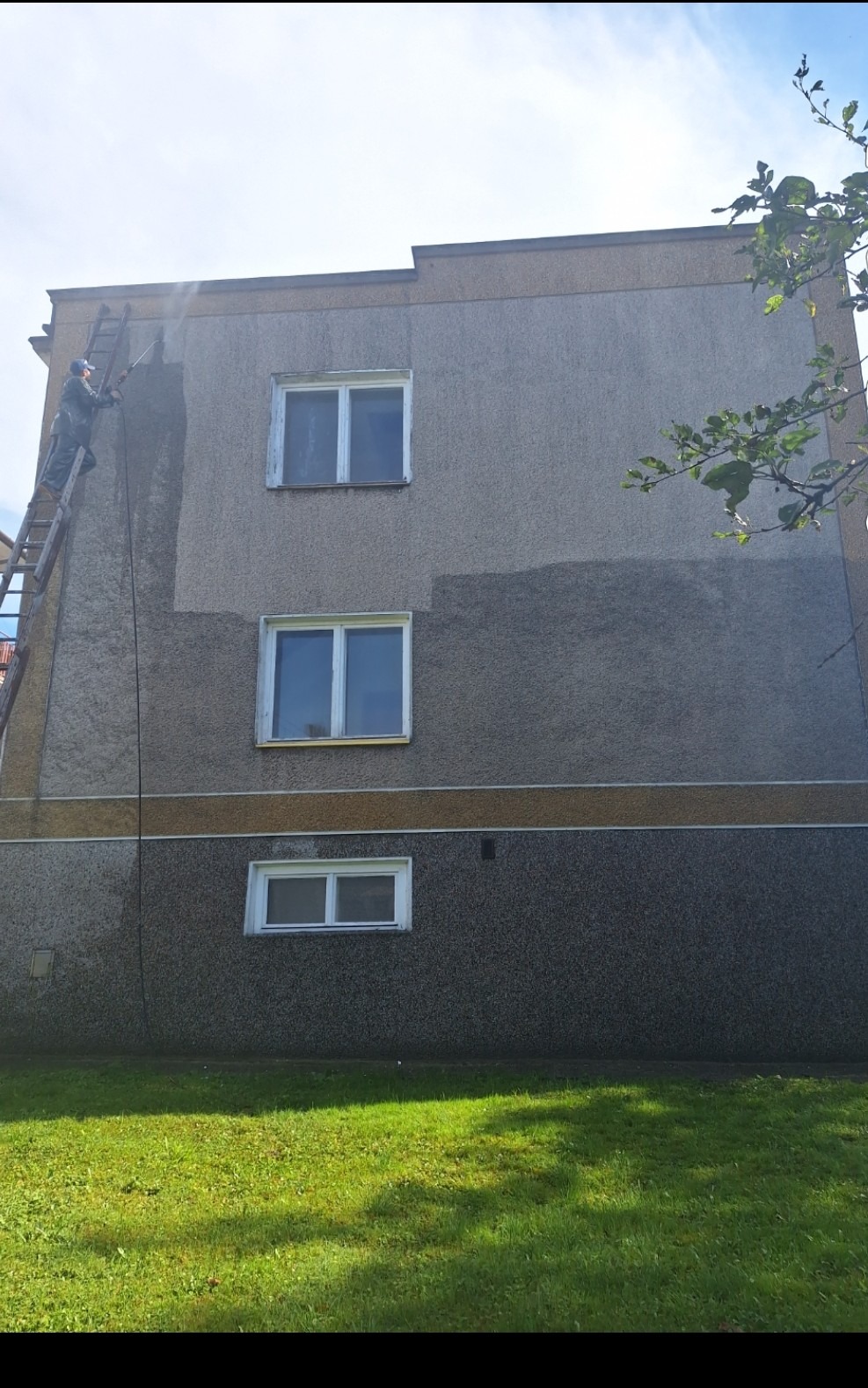 Pracownik na drabinie spryskuje elewację budynku, widoczna różnica w kolorze po umyciu fragmentu ściany, trzy okna na różnych poziomach, trawnik u dołu.