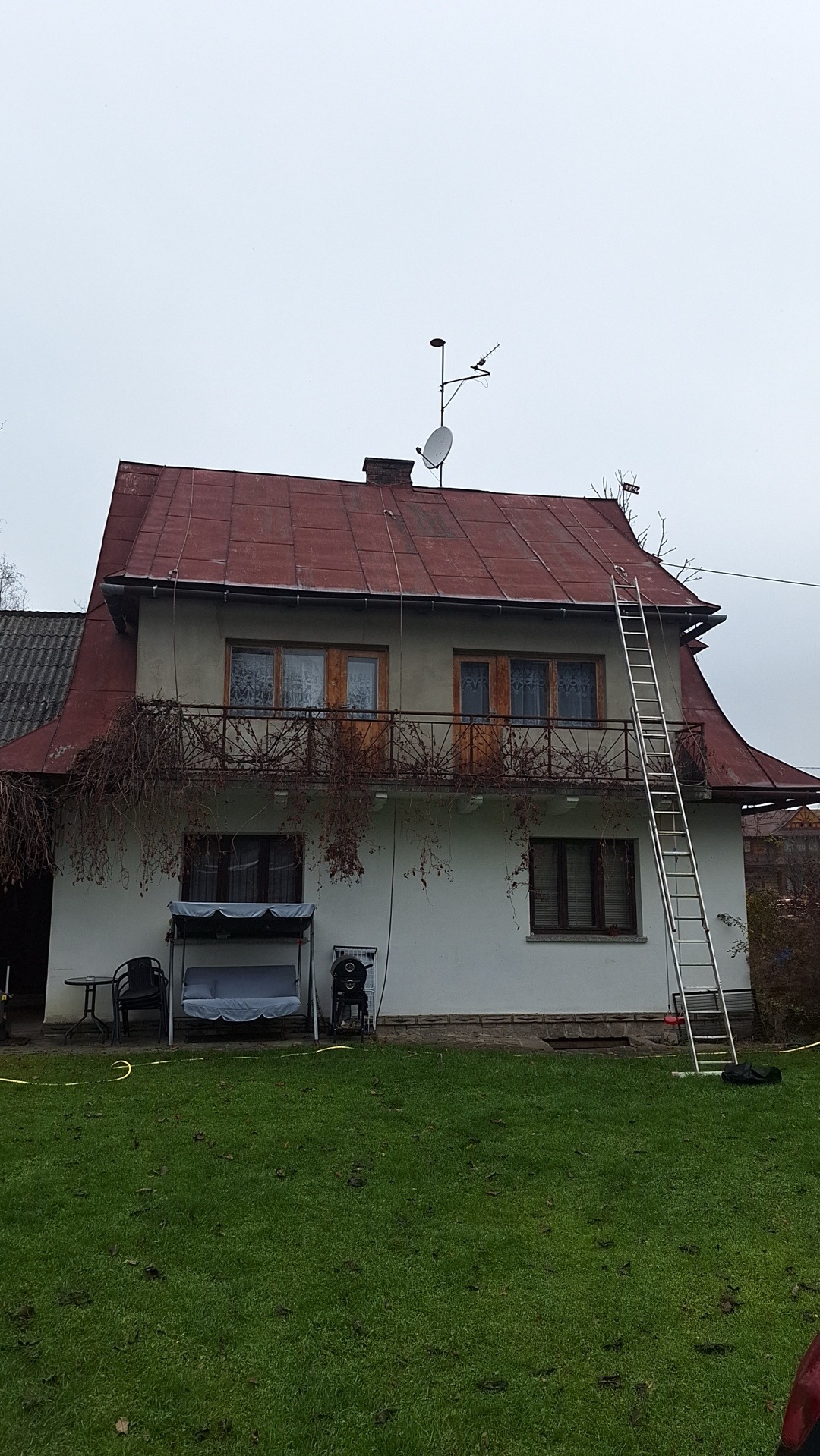 Dwupiętrowy dom z czerwonym blaszanym dachem, balkonem porośniętym suchymi pnączami i przystawioną aluminiową drabiną.