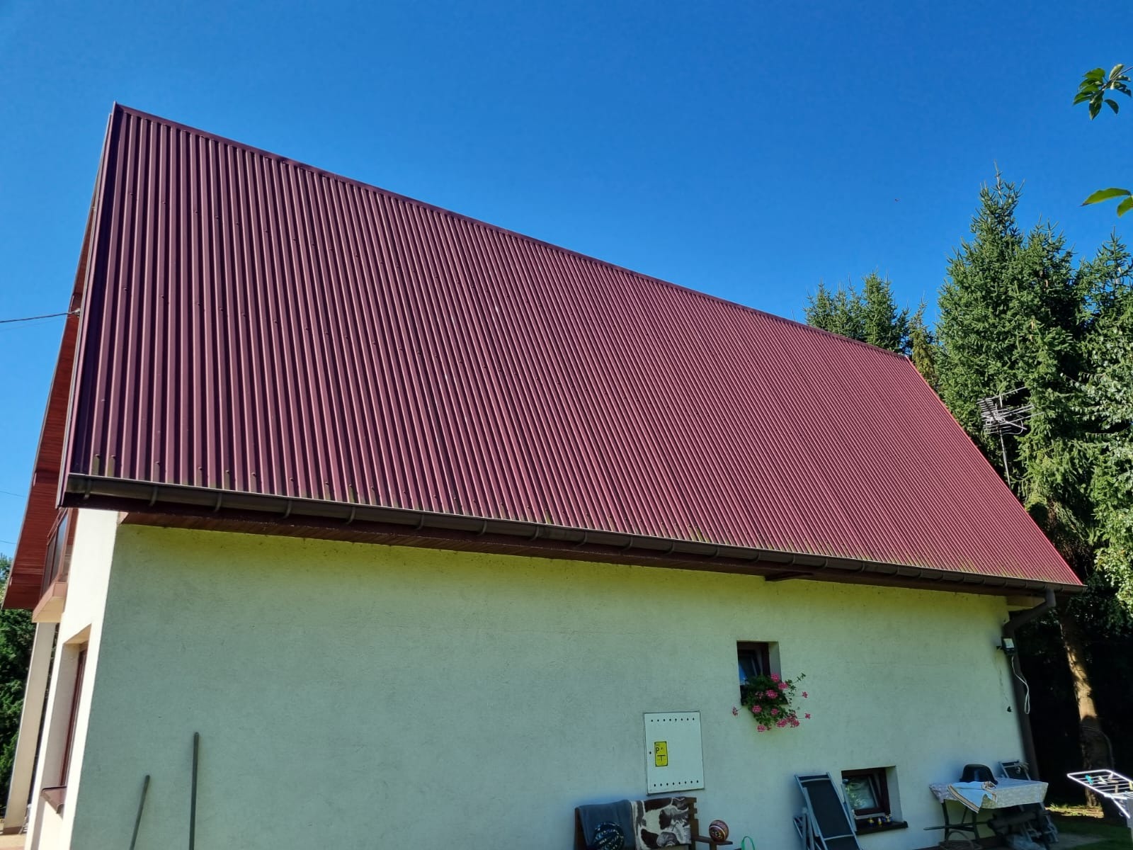 Dom jednorodzinny z nowym, bordowym dachem z blachy trapezowej, widok z dołu na tle błękitnego nieba. Dach wykończony brązowymi rynnami. Na ścianie domu skrzynka elektryczna i okno z kwiatami.
