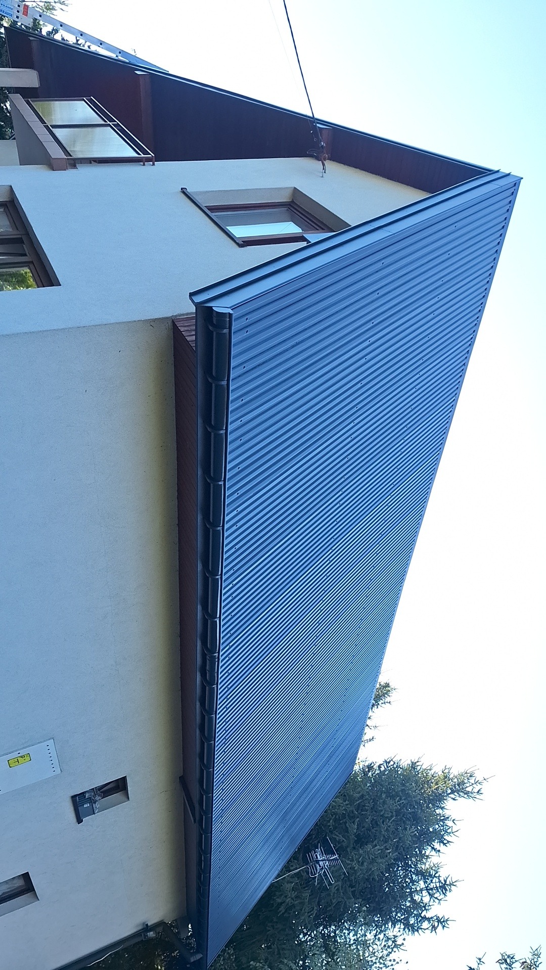 Fragment elewacji budynku z nową, niebieską blachą trapezową, widok z dołu, z widocznym fragmentem dachu i drzewa w tle.