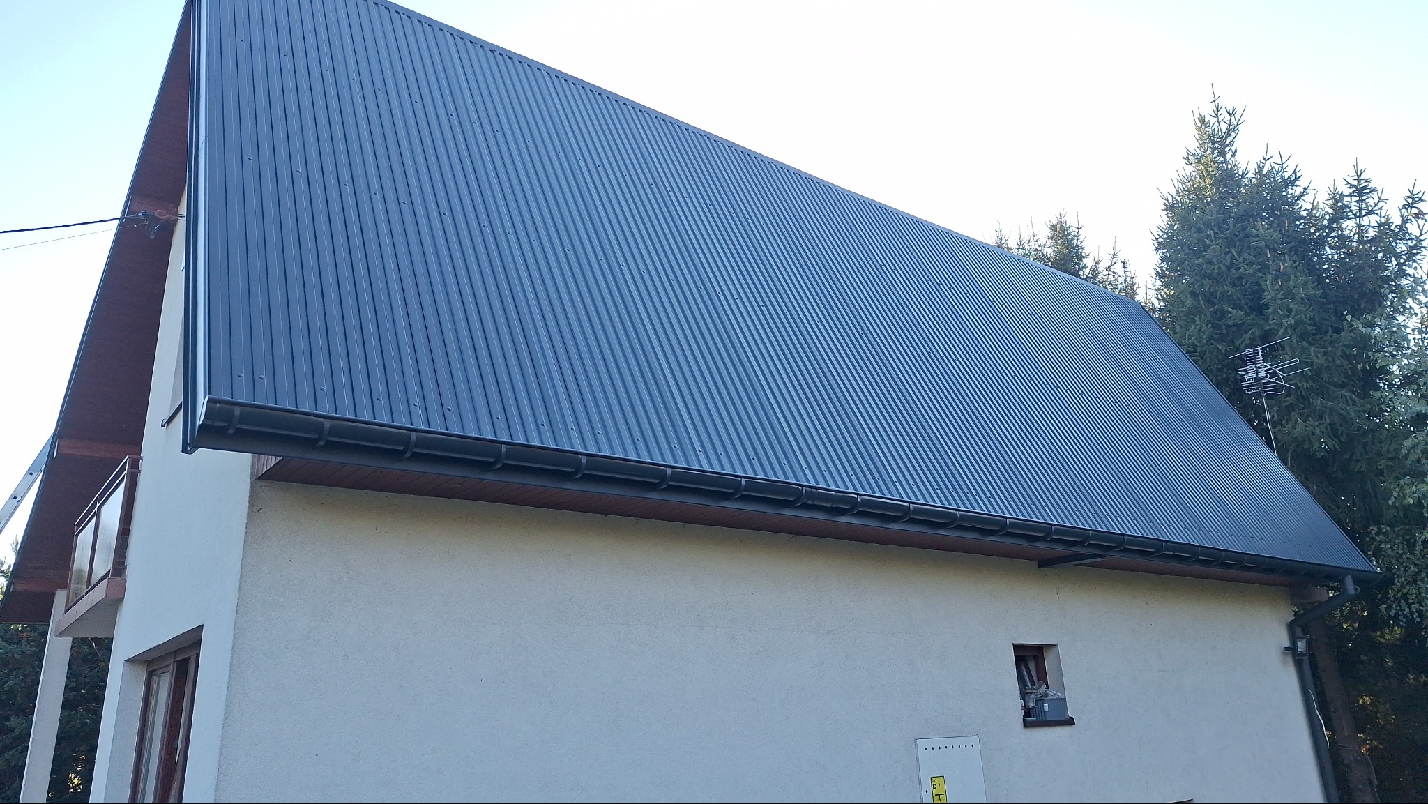 Dom jednorodzinny z nowym, ciemnoszarym dachem z blachy trapezowej i zamontowanymi rynnami. Widoczna elewacja w jasnym kolorze. W tle iglaste drzewa.