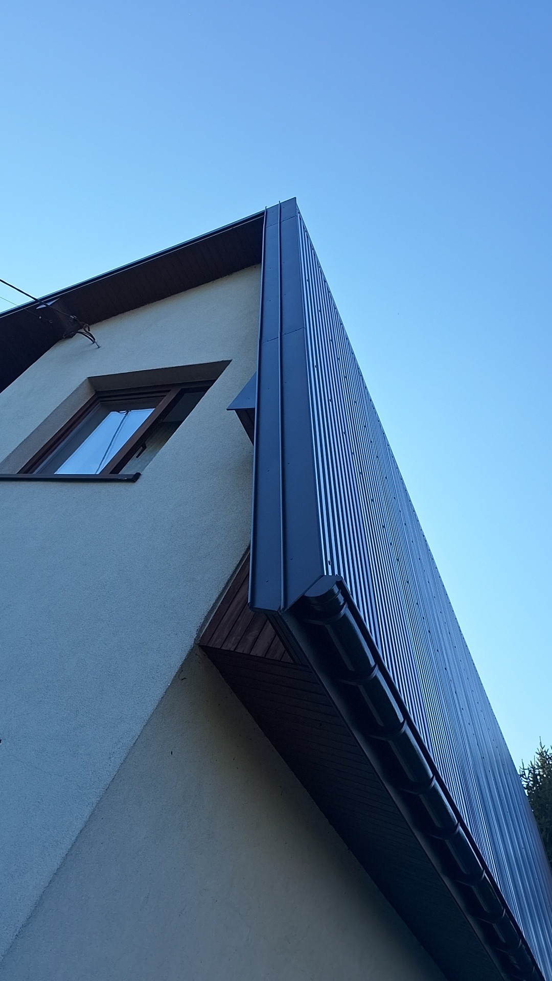 Fragment elewacji budynku z nowoczesnym, pionowo żłobkowanym, ciemnoszarym panelem elewacyjnym, widok z dołu na tle błękitnego nieba.