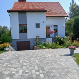 Dwukondygnacyjny dom z czerwonym dachem i jasną elewacją, ozdobiony kwiatami w donicach i rabatami kwiatowymi, z widoczną kostką brukową na podjeździe.