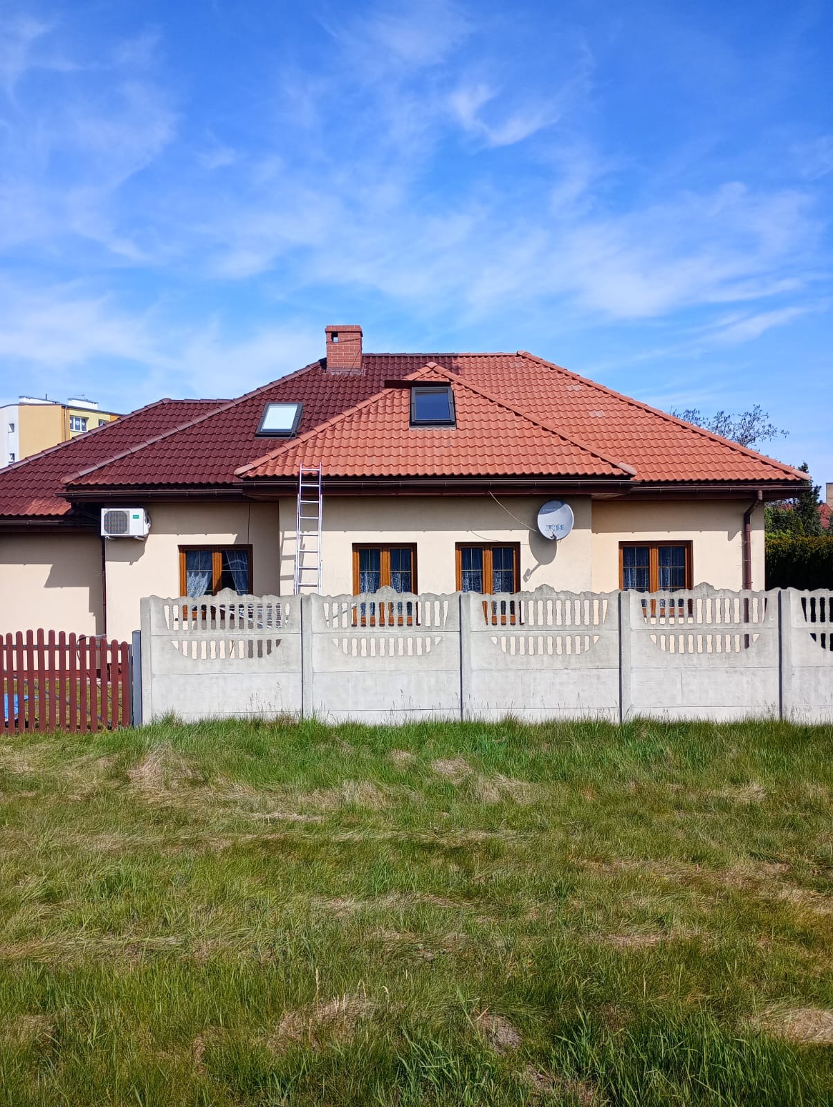 Dom jednorodzinny z częściowo odnowionym dachem w odcieniach czerwieni, jasnobeżową elewacją i betonowym ogrodzeniem, widziany z perspektywy trawnika.