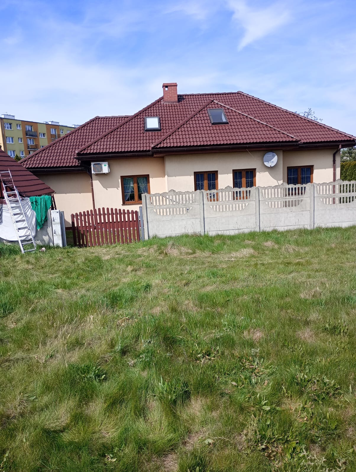 Dom jednorodzinny z brązowym dachem i beżową elewacją, widoczna drabina oparta o ścianę, trawnik z przodu, betonowe i drewniane ogrodzenie.