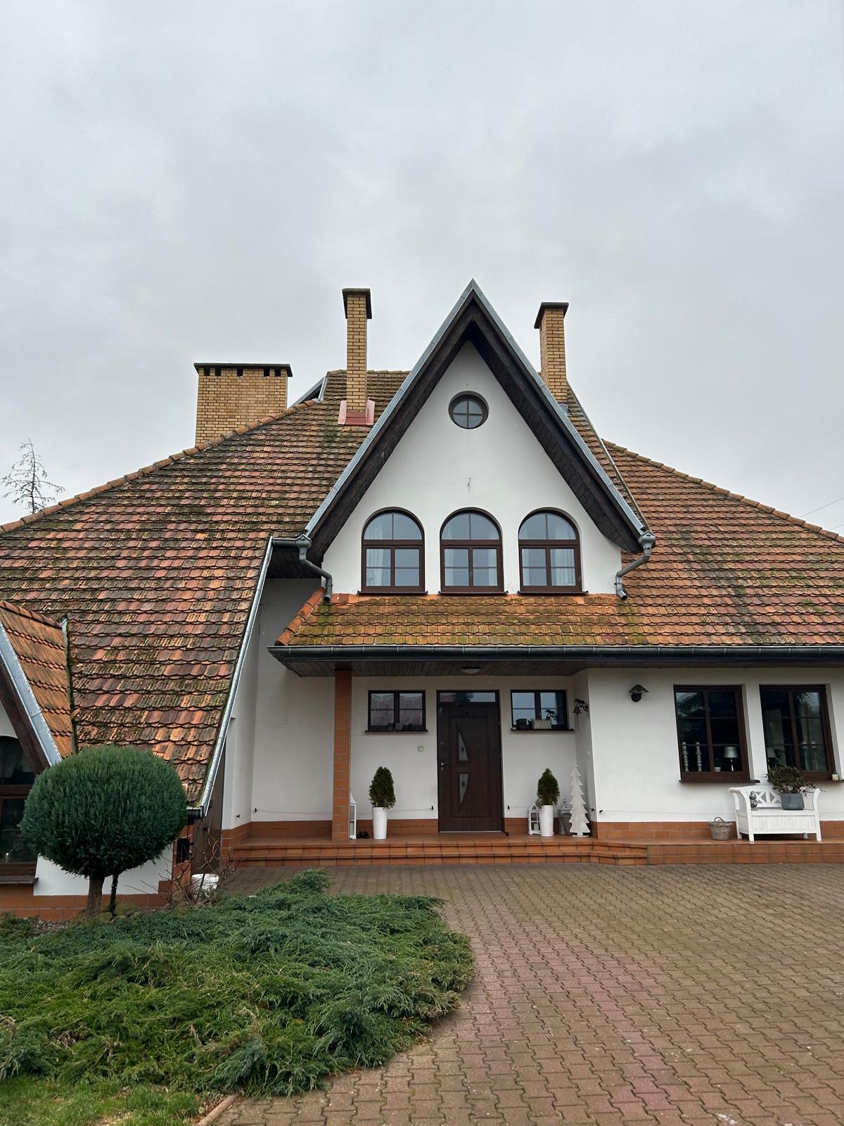 Dwupiętrowy dom z czerwoną dachówką, widoczne dwa kominy, trzy okna łukowe na górze i biała elewacja, na podwórku rosną krzewy
