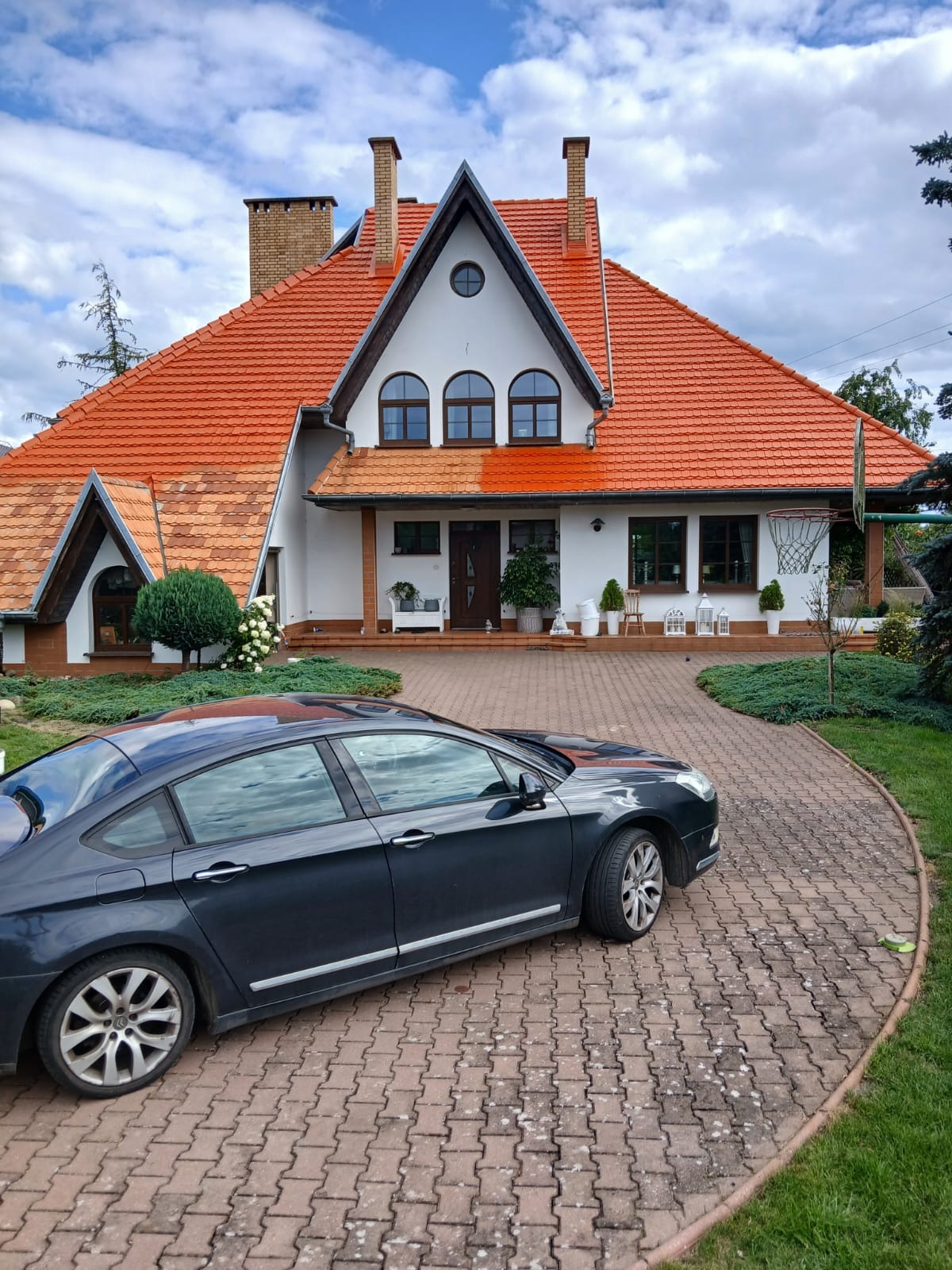 Dom jednorodzinny z częściowo umytym dachem z czerwonej dachówki, widoczna różnica w kolorze między czystą a brudną powierzchnią. Na podjeździe stoi ciemny samochód.