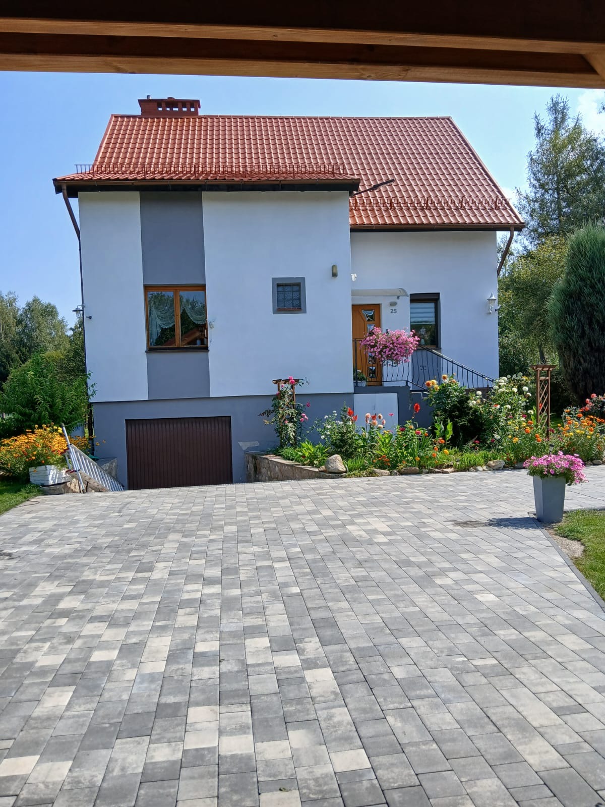 Dwukondygnacyjny dom z czerwonym dachem i jasną elewacją, ozdobiony kwiatami w donicach i rabatami kwiatowymi, z widoczną kostką brukową na podjeździe.