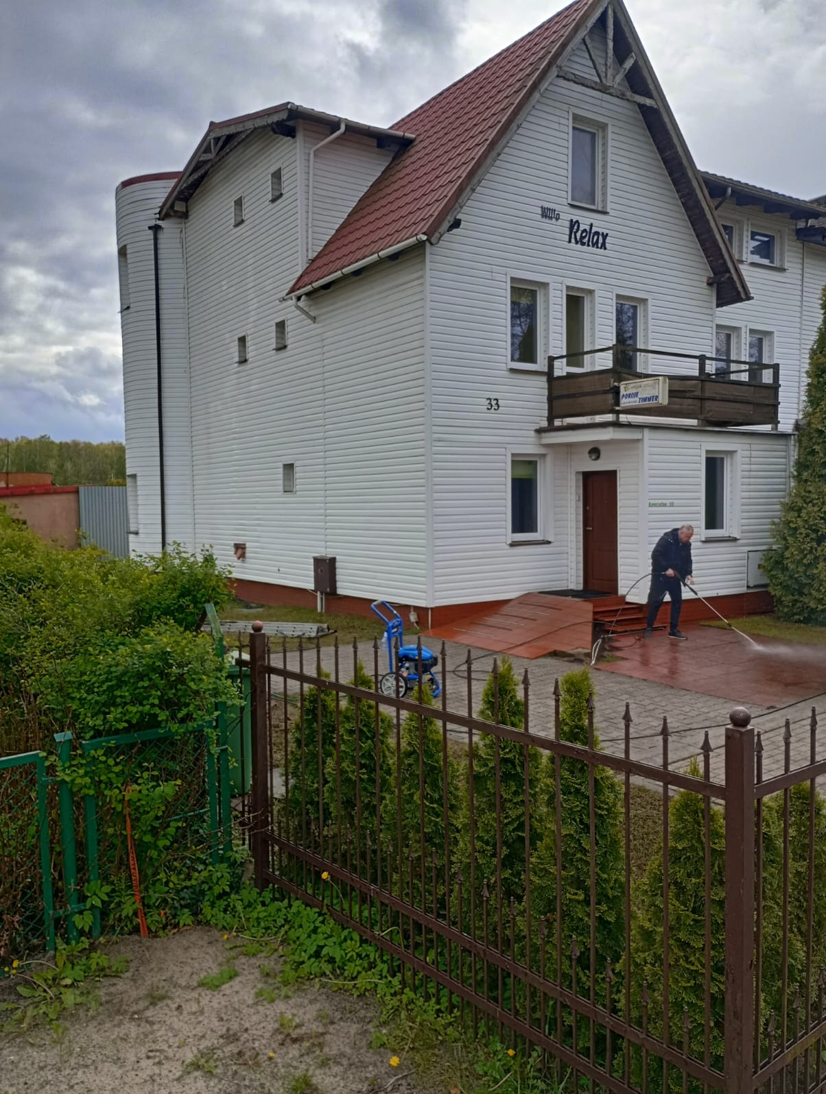 Pracownik w ciemnej odzieży czyści ciśnieniowo chodnik przed budynkiem z białym sidingiem i czerwoną dachówką; widoczny niebieski agregat ciśnieniowy i metalowe ogrodzenie.