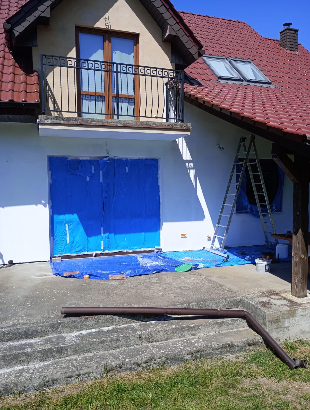Przygotowanie do malowania elewacji domu: okna i drzwi zabezpieczone folią, drabina oparta o ścianę, widoczne wiadro z farbą i rozłożona folia ochronna na ziemi.