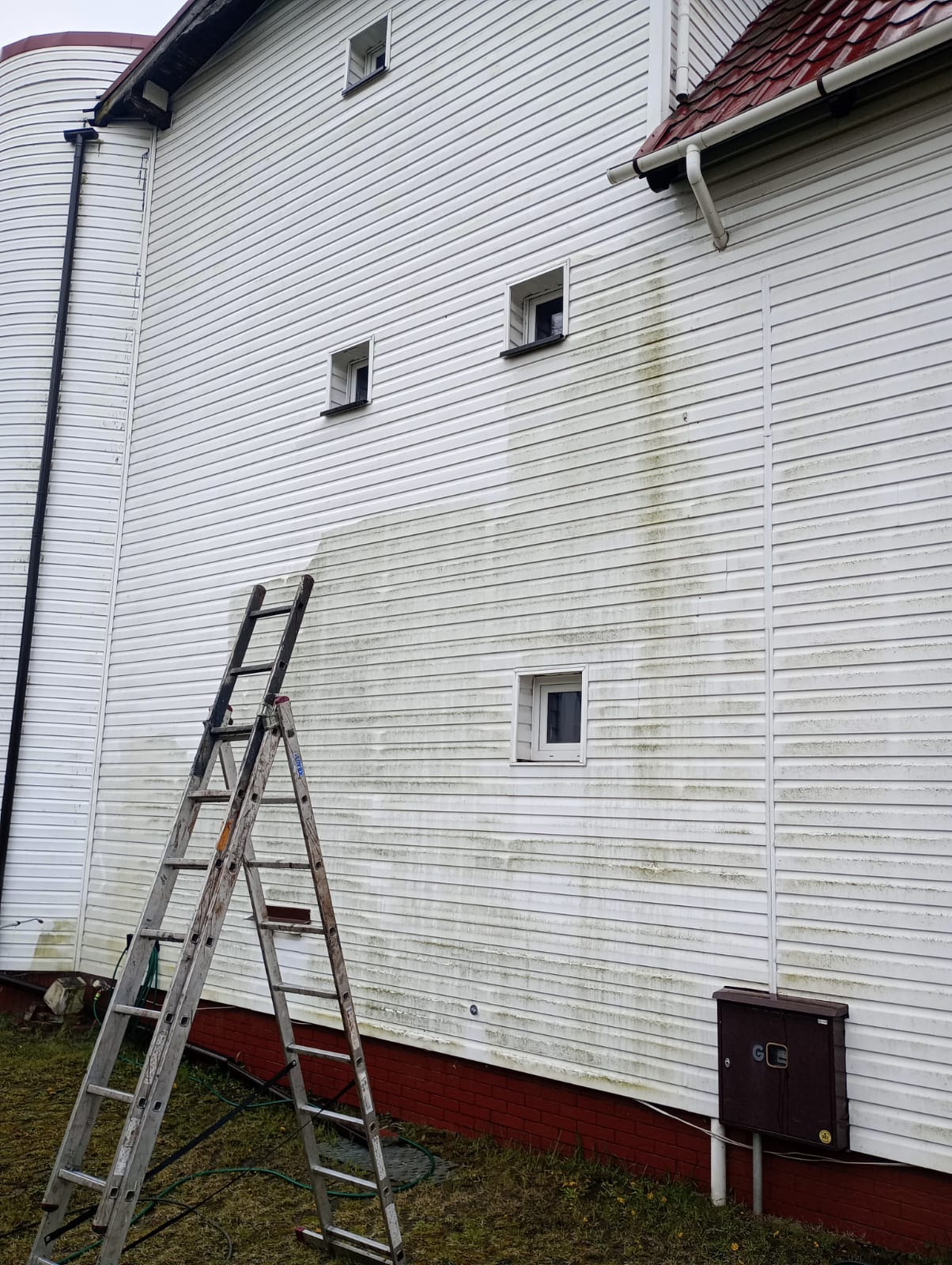 Fragment ściany budynku z białym sidingiem, częściowo wyczyszczonym z zabrudzeń, z widoczną drabiną opartą o ścianę.