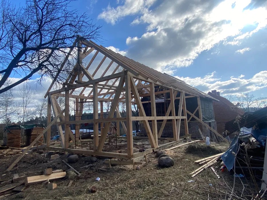 Konstrukcja szkieletowa domu z drewna w trakcie budowy, widoczna więźba dachowa i elementy ścian, w tle budynek z czerwonej cegły i błękitne niebo z chmurami.